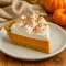 Slice of no-bake pumpkin pie with a creamy pumpkin-cream filling and fluffy marshmallow topping in a graham cracker crust, served on a white plate with a sprinkle of pumpkin spice.