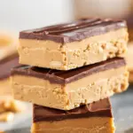 Chocolate-drizzled peanut butter protein bars cut into neat rectangles on parchment, with a spoonful of peanut butter and a scattering of oats nearby.