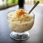 Small glass of silky coconut pudding topped with toasted coconut flakes and a lime twist, served on a rustic plate.