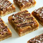 Stack of caramel coconut bars with toasted coconut and chocolate drizzle on parchment-lined board.