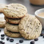 Stack of round espresso-glazed sugar cookies on a wooden board, with scattered coffee beans and a drizzle of glaze.