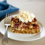 A slice of Healthy Pumpkin Dump Cake with a golden, crunchy pecan topping served on a rustic white plate. The moist, custardy pumpkin layer glows beneath the crumbly topping, dusted lightly with cinnamon sugar. A dollop of whipped cream melts over the warm Gluten Free Pumpkin Cake, surrounded by autumn leaves and a cup of coffee for the perfect Easy Gluten Free Desserts fall moment.