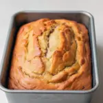 Close-up of a moist slice of banana bread studded with chopped walnuts on a wooden board, a second loaf visible in the background.