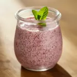 Jar of pink strawberry chia pudding topped with sliced strawberries and a mint leaf, spoon at the ready.