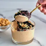 Close-up of a creamy peanut-butter mousse in a small jar, drizzled with melted chocolate and sprinkled with chopped peanuts, spoon on the side.