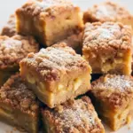 A golden, moist Gluten Free Apple Cake topped with cinnamon sugar, sliced to reveal tender apple layers — the perfect fall-inspired Gluten Free Sweets dessert served on a rustic wooden table with apples and cinnamon sticks nearby.