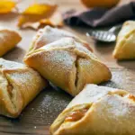 Three golden pumpkin cream cheese danishes on a wooden board, showing flaky puff pastry, a creamy pumpkin-cream center, and a light dusting of powdered sugar.