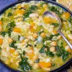 Steaming bowl of orzo vegetable soup with chickpeas, kale, lemon wedge, and a slice of crusty bread on a wooden board.