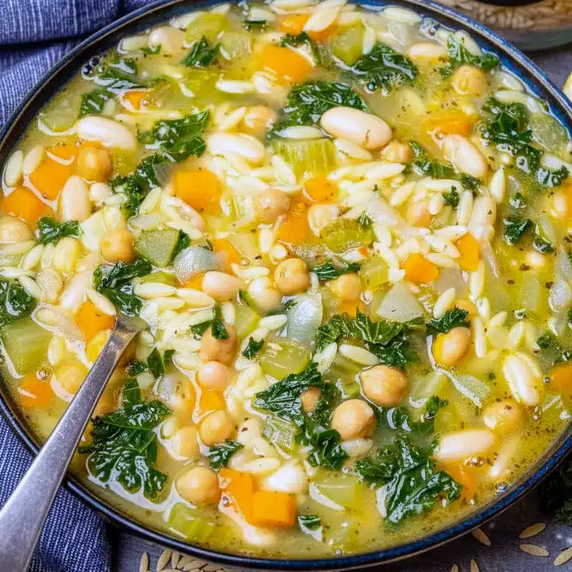 Steaming bowl of orzo vegetable soup with chickpeas, kale, lemon wedge, and a slice of crusty bread on a wooden board, Vegan Soup Recipes.