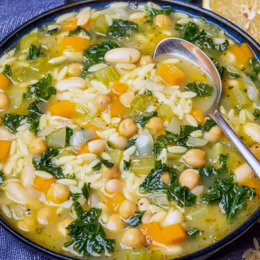 Steaming bowl of orzo vegetable soup with chickpeas, kale, lemon wedge, and a slice of crusty bread on a wooden board.