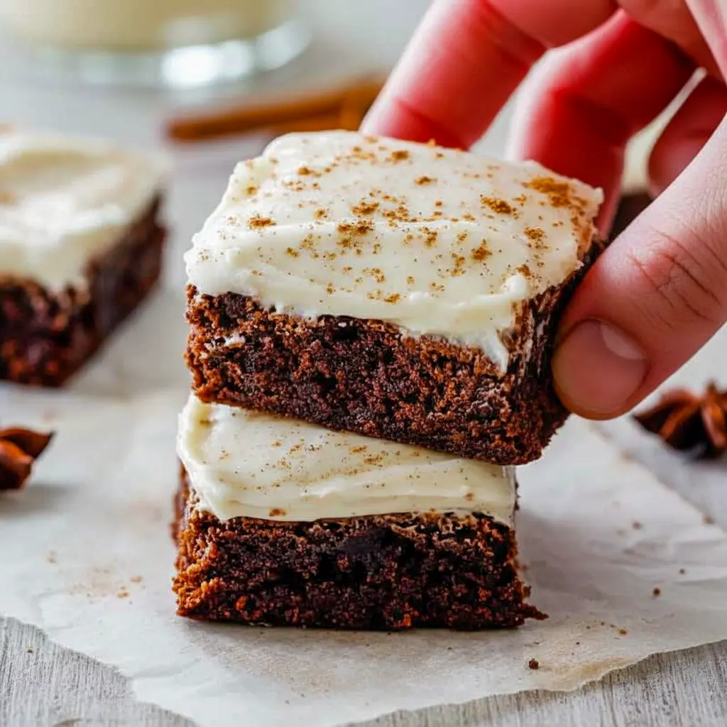A platter of square gingerbread brownies frosted with creamy eggnog cream-cheese icing, dusted with cinnamon and arranged on a festive holiday napkin, Gingerbread Brownies With Eggnog Frosting.