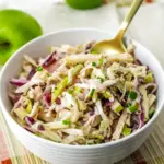 Close-up of a white bowl filled with crisp apple coleslaw: shredded cabbage, matchstick apples, celery, and green onions tossed in a warm apple-cider dressing, ready for serving.