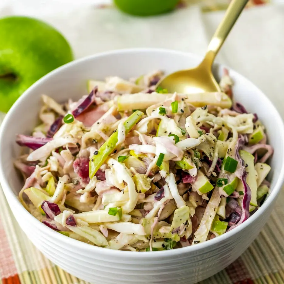 Close-up of a white bowl filled with crisp apple coleslaw: shredded cabbage, matchstick apples, celery, and green onions tossed in a warm apple-cider dressing, ready for serving.