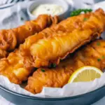 Golden fillets with a super-crispy coating on a wire rack, served with lemon wedges and a bowl of tartar sauce.