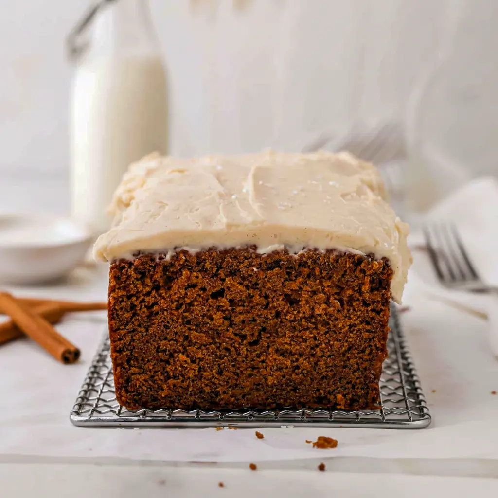Slice of moist ginger loaf with brown-butter cream cheese frosting on a wooden board, dusted with cinnamon and styled with a small rosemary sprig, Ginger Loaf Bread.