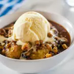 Skillet of golden banana crumble with oat-pecan streusel, a melting scoop of vanilla ice cream and a light caramel drizzle, served on a white plate.
