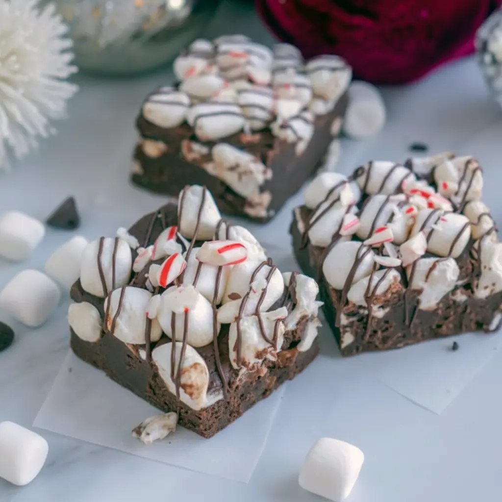 Close-up of peppermint chocolate fudge squares topped with mini marshmallows, crushed candy cane, and a chocolate drizzle, arranged on a festive napkin, Peppermint Chocolate Fudge.