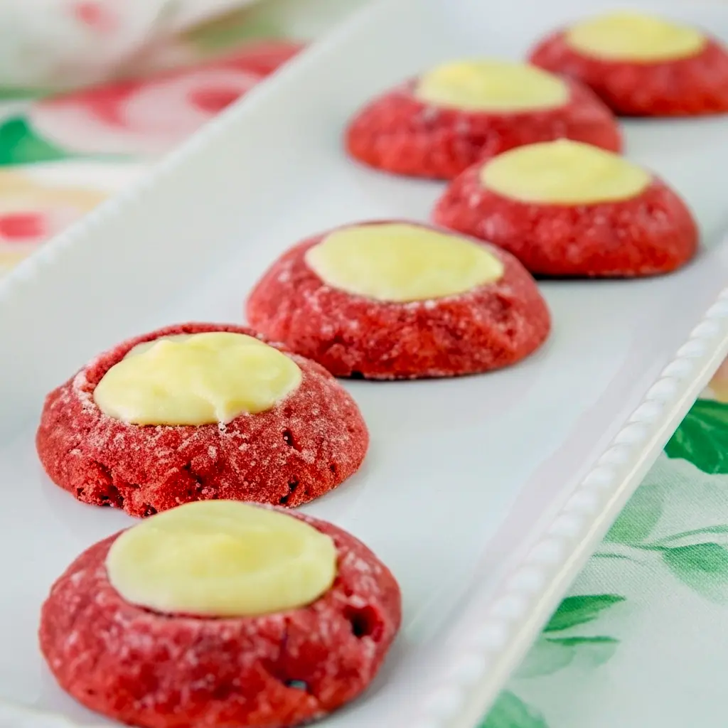 Close-up of glossy red velvet thumbprint cookies filled with creamy white cream cheese, arranged on a festive plate with a light dusting of powdered sugar and red sanding sugar, Best Xmas Cookie Recipes.