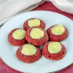 Close-up of glossy red velvet thumbprint cookies filled with creamy white cream cheese, arranged on a festive plate with a light dusting of powdered sugar and red sanding sugar, Best Xmas Cookie Recipes.
