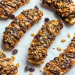 Frozen banana slices topped with peanut butter, chopped peanuts, and dark chocolate drizzle on parchment paper, Healthy Banana Treats.