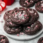 Chocolate Peppermint Christmas Cookies — stack of glossy double-chocolate cookies dotted with peppermint chips and crushed candy cane on a parchment-lined tray.