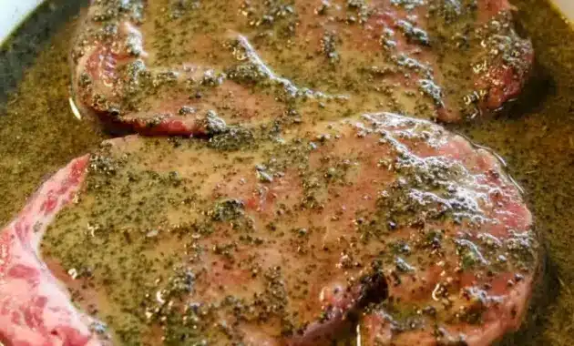 Jar of Steak Marinade beside raw steaks on a cutting board, with fresh herbs, lemon, and a small bowl of sauce ready for brushing on the grill.