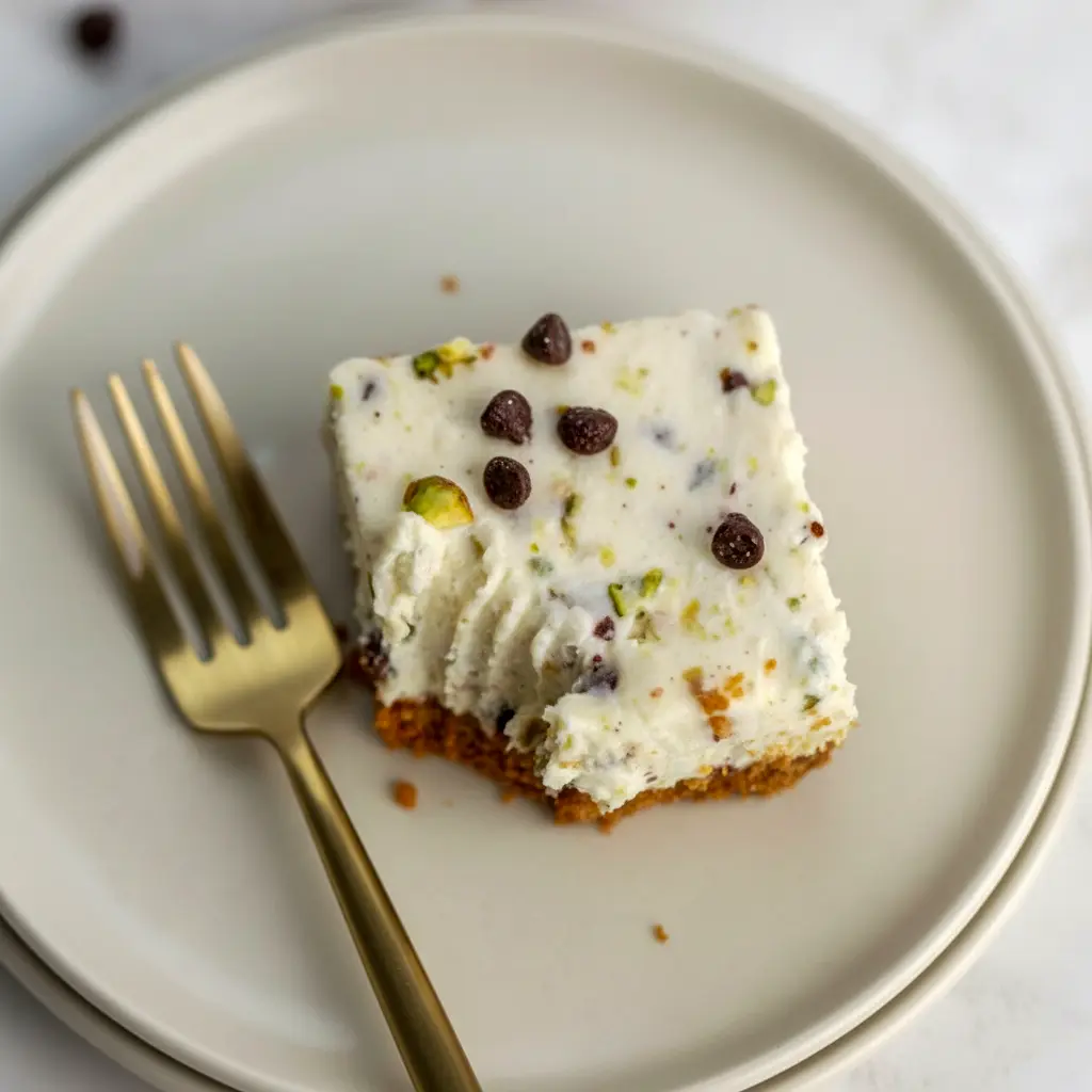 Square no-bake cannoli cheesecake bars on a serving plate, topped with mini chocolate chips and chopped pistachios.