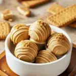 Close-up of round white-chocolate–coated truffles with cookie crumb garnish on parchment paper, showing glossy drizzle and warm cinnamon specks, No Bake Biscoff Truffles.