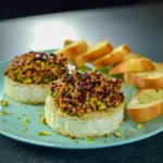 Small Group Appetizers — golden baked brie topped with chopped pistachios and honey, on a wooden board with toasted baguette rounds.