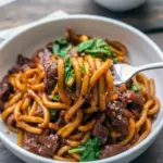 Close-up of black pepper beef udon: glossy noodles with sliced beef, onions, and cabbage coated in a dark pepper sauce, topped with chopped green onions, Asian Recipes.