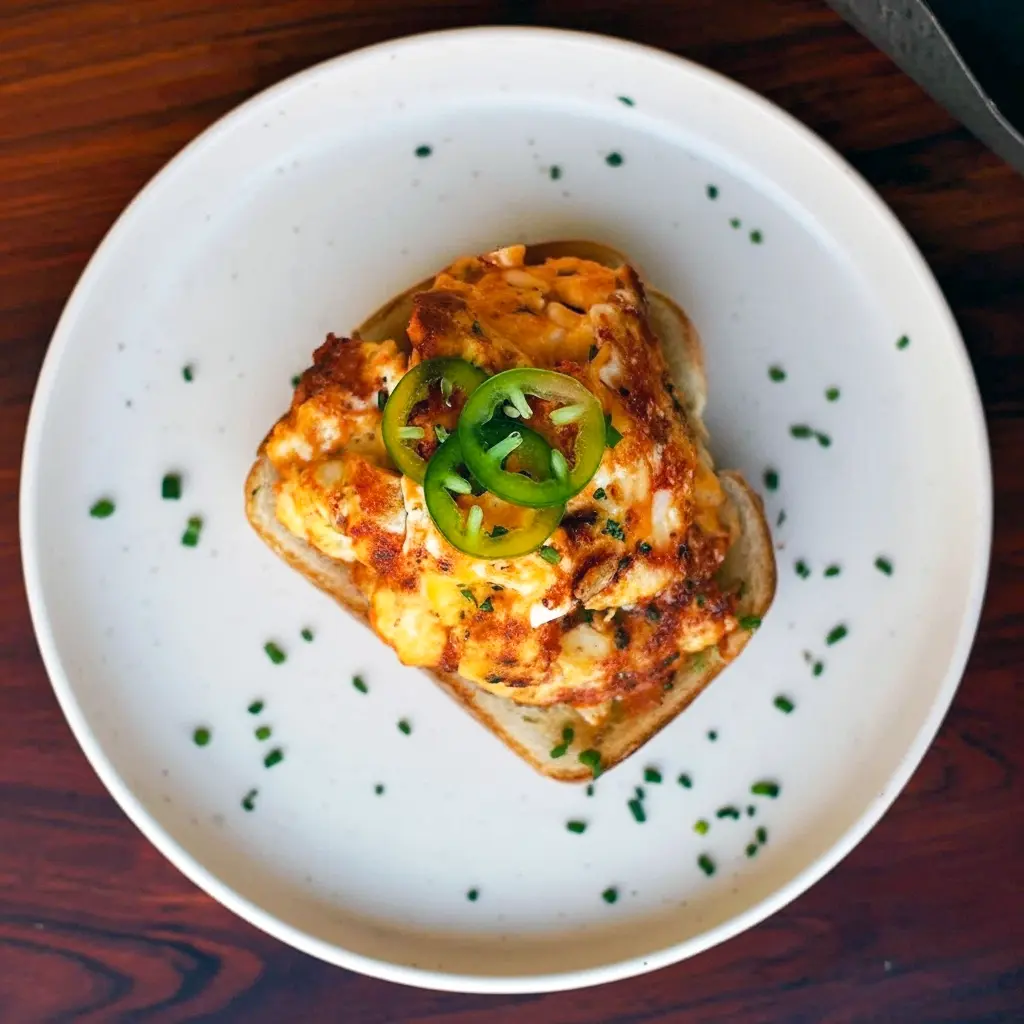 Golden-brown egg salad cakes stacked on toasted sourdough with mashed avocado and jalapeño slices, skillet in the background, Crispy Egg Salad.