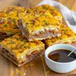 Stacked golden French Dip Squares on a white plate with melted cheese and caramelized onions, a small bowl of steaming au jus for dipping in the background, Holiday Dinner Inspiration.