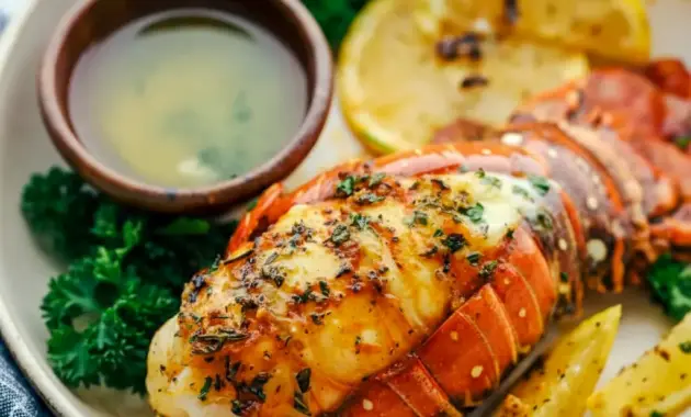 Broiled Lobster Tail Recipe topped with garlic-herb butter, lemon wedge, and chopped parsley on a serving plate.