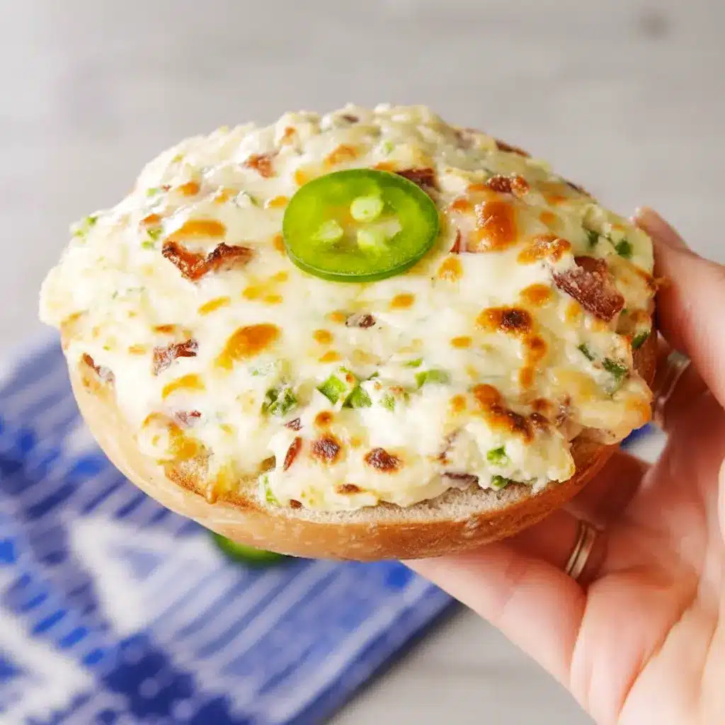 Close-up of baked Jalapeño Popper Bagels — bagel halves spread with jalapeño cream cheese, melted Monterey Jack, and bacon crumbles, garnished with thin jalapeño slices.