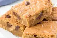 A close-up tray of Peanut Butter Blondies topped with Peanut Blondies style texture, loaded with Reese’s Pieces and chocolate chips, showing soft centers and chewy edges.